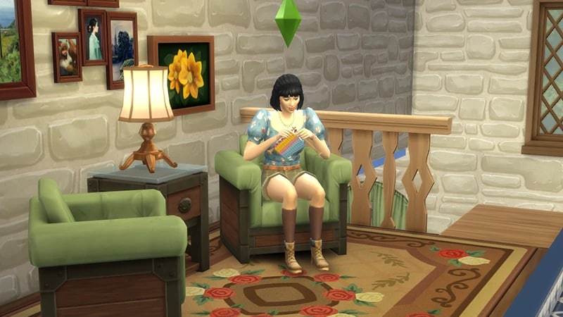 A Sim with dark hair sits on a green armchair, in her hands are two needles as she focuses on knitting something.