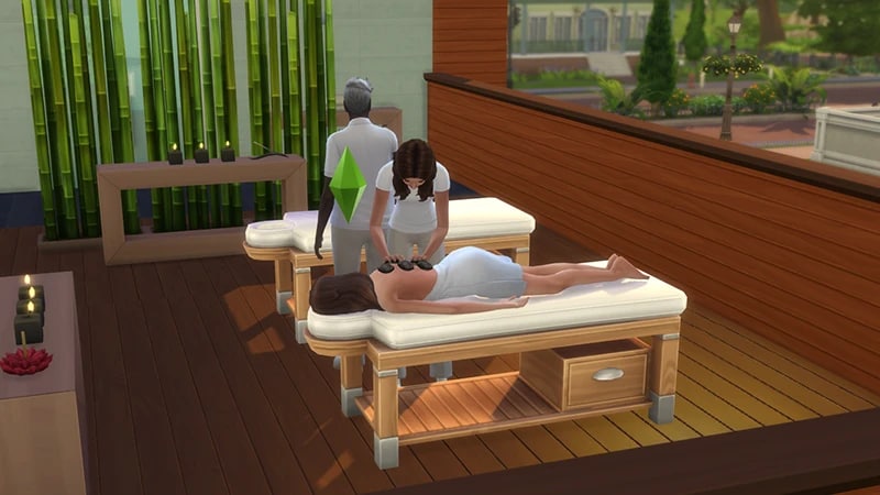 Bir Sim krem rengi ahşap bir masaj yatağında uzanırken, beyaz giyen bir Sim de onun sırtına sıcak taşlar yerleştiriyor.