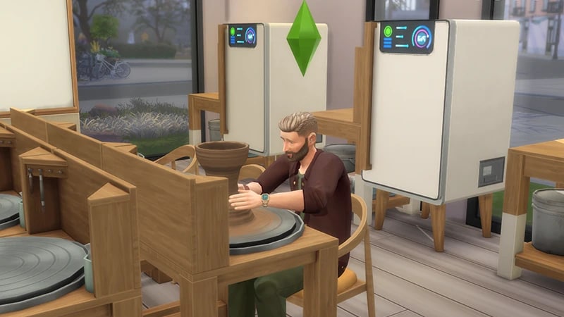 A bearded Sim sits at a pottery wheel near a window in a pottery studio crafting a vase from clay.