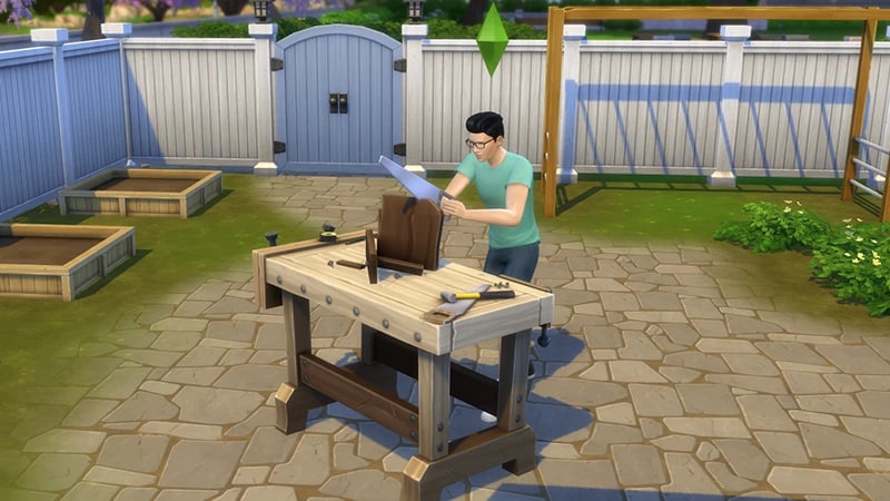 A Sim with black hair, a green t-shirt, and glasses, stands in a garden sawing a piece of wood at the woodworking table.